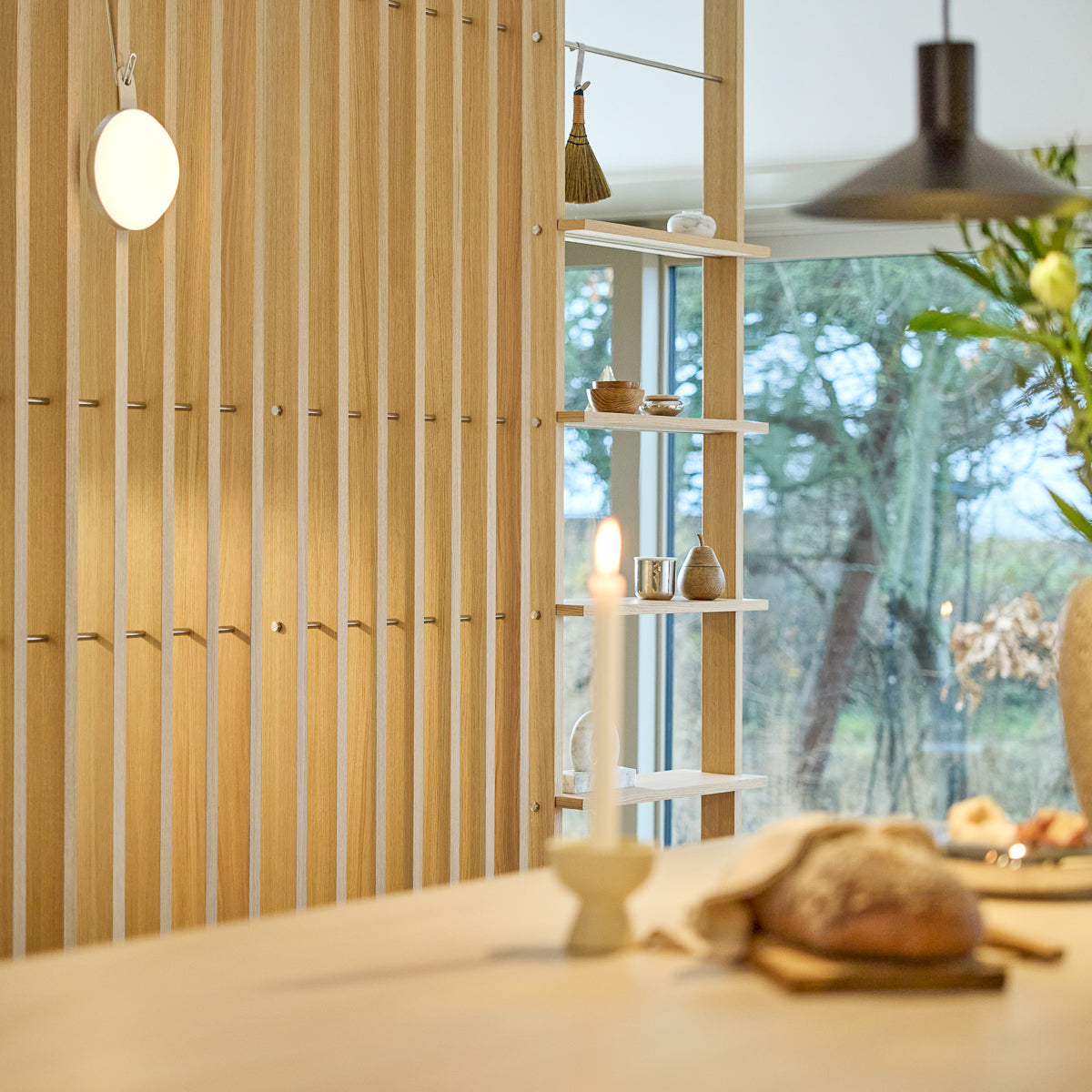Minimalistisch interieur met een houten ruimteverdeler Nordic EMBRACE, brandende kaars, vers brood op tafel, verzorgde decoratie op de planken en raam dat buiten bomen omlijst.